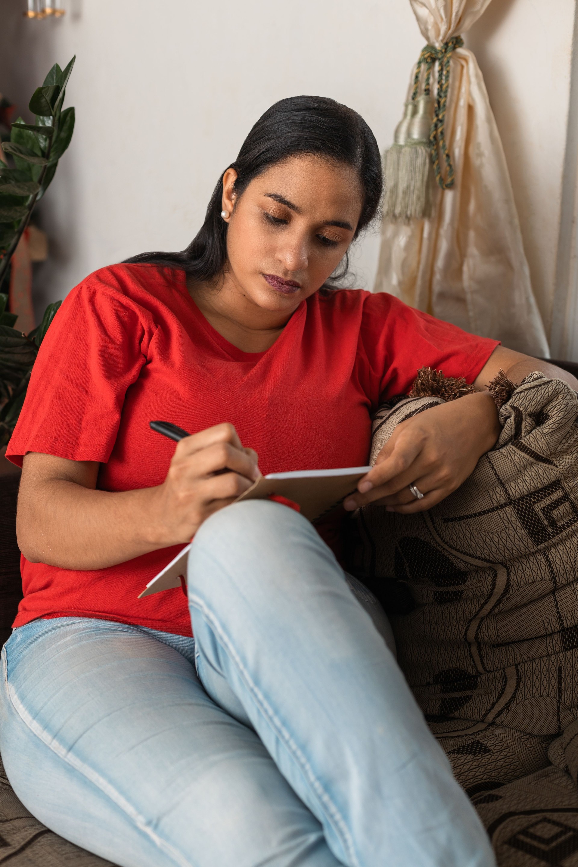 mujer latina irreconocible, sentada en el sofá escribiendo en un cuaderno, suéter rojo, jeans azules, tiro vertical,